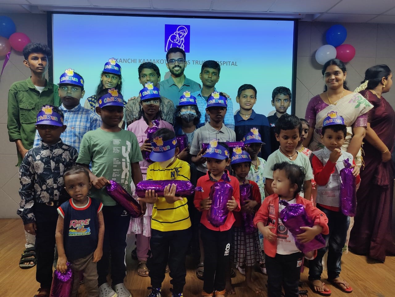Children in blue hats holding gifts, with adults and a screen behind.