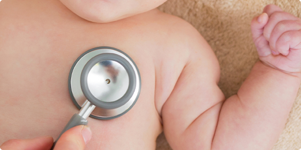 A Medical Professional's Hand Holds a Stethoscope to a Baby's Chest.