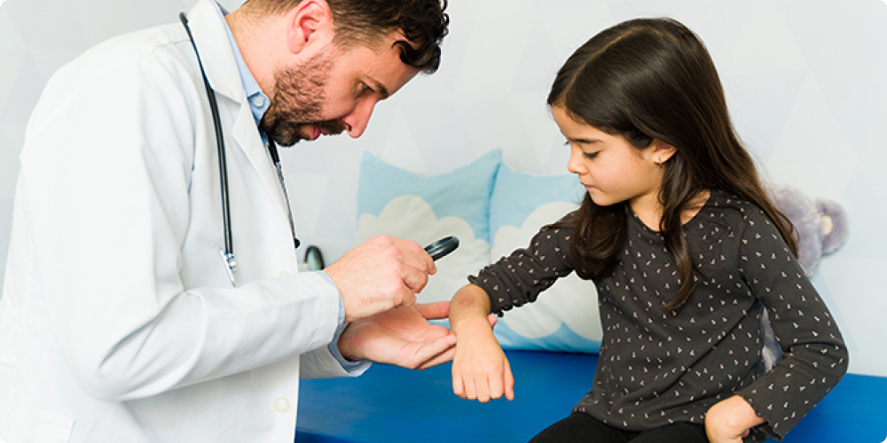 A male doctor in a white lab coat examines a young girl's arm with a magnifying tool.